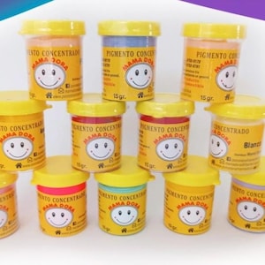 May include: A collection of 11 small, yellow plastic jars with lids. Each jar contains a concentrated pigment and has the text "PIGMENTO CONCENTRADO MAMA DORA" printed on the label. The jars are arranged in a circle on a white surface.
