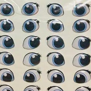 May include: A sheet of 24 googly eyes stickers. The eyes are white with black pupils and blue irises. The stickers are arranged in four rows of six. The sheet is labeled "Art 076 Mediano".