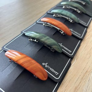 May include: A series of six curved hair barrettes with smooth, polished finishes in shades of orange, dark green, and light green. Each barrette is attached to a black rectangular display card featuring the word Accessory in white script text.