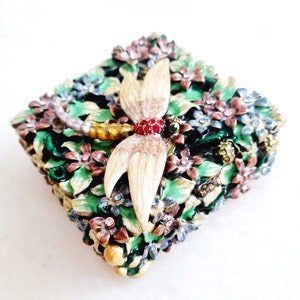 May include: A square trinket box with a detailed design of flowers and a dragonfly. The dragonfly is made of metal and has a light brown body with white wings. The box is decorated with green, blue, pink, and gold accents.