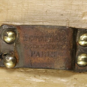 May include: A close-up of a brown leather tag with the text "E.Goyardine Gare St. Lazare Paris" embossed on it. The tag is attached to a wooden surface with four gold-coloured rivets.
