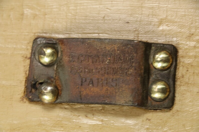 May include: A close-up of a brown leather tag with the text "E.Goyardine Gare St. Lazare Paris" embossed on it. The tag is attached to a wooden surface with four gold-coloured rivets.