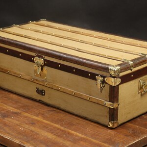 May include: A vintage, tan canvas luggage trunk with brown leather trim and brass hardware. The trunk has a rectangular shape and is sitting on a wooden surface.