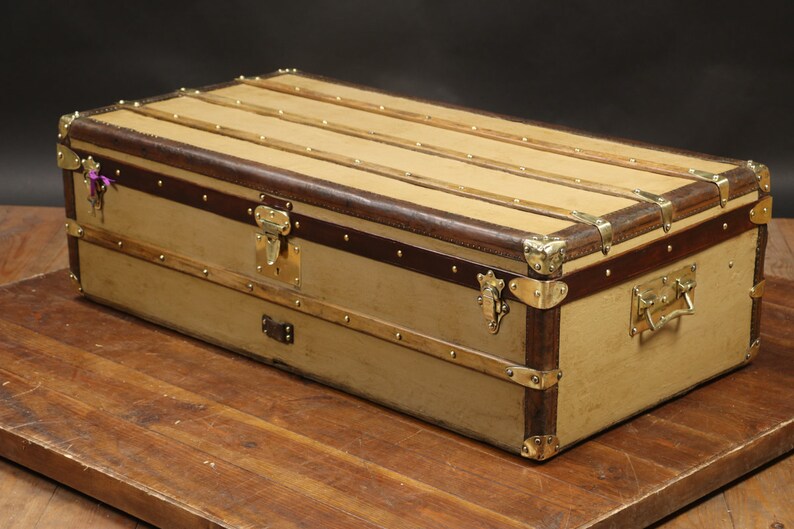 May include: A vintage, tan canvas luggage trunk with brown leather trim and brass hardware. The trunk has a rectangular shape and is sitting on a wooden surface.
