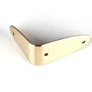 May include: Gold-colored metal guitar strap button with two holes. The button is shaped like a rounded triangle.