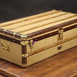 May include: A vintage, tan canvas travel trunk with brown leather trim and brass hardware. The trunk has a hinged lid with a brass lock and a handle on the front.