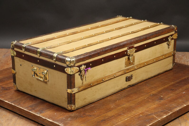 May include: A vintage, tan canvas travel trunk with brown leather trim and brass hardware. The trunk has a hinged lid with a brass lock and a handle on the front.
