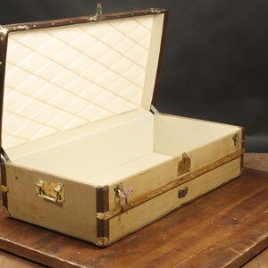 May include: A vintage-style, tan canvas travel trunk with brass hardware and a brown leather trim. The trunk is open and the interior is lined with a white quilted fabric.