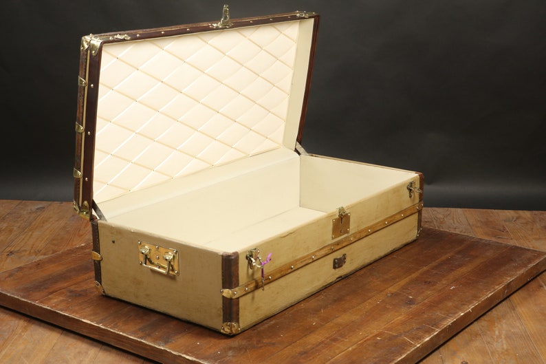 May include: A vintage-style, tan canvas travel trunk with brass hardware and a brown leather trim. The trunk is open and the interior is lined with a white quilted fabric.