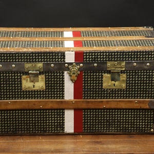 Op de afbeelding: Een vintage reiskoffer met een zwart-wit geruit patroon en een rode en witte streep in het midden. De koffer heeft messing beslag en een houten frame.