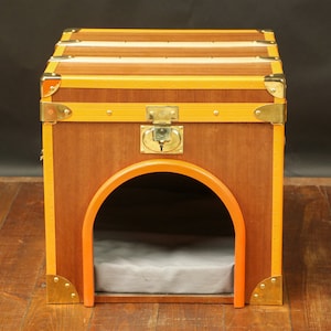 Peut inclure: Une niche pour chien en bois en forme de valise vintage avec une serrure en laiton et une bordure jaune et orange. La niche a un coussin gris à l'intérieur et une grande ouverture pour que le chien puisse entrer.