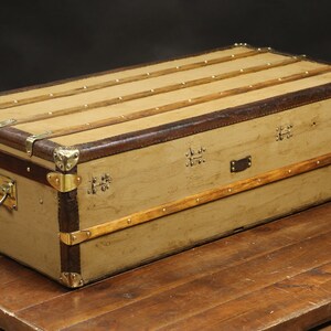 May include: A vintage canvas and leather travel trunk with brass hardware and wooden accents. The trunk has a rectangular shape and is closed.