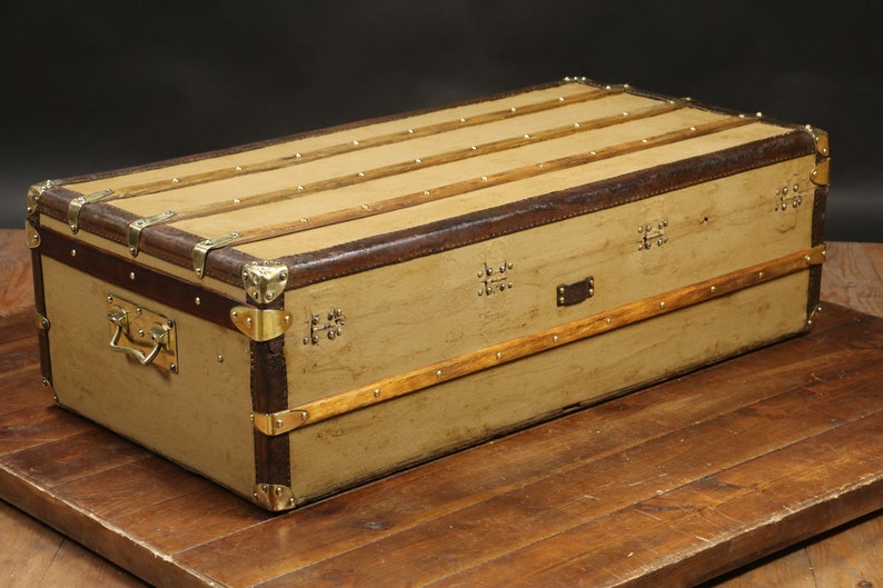 May include: A vintage canvas and leather travel trunk with brass hardware and wooden accents. The trunk has a rectangular shape and is closed.