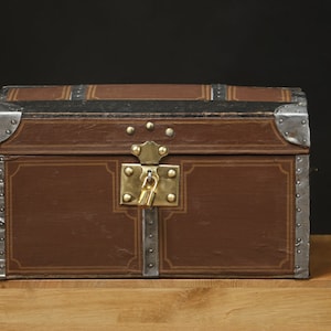 Puede incluir: Un cofre de madera marrón con detalles metálicos y una cerradura de latón. El cofre tiene un aspecto desgastado y vintage y está decorado con un diseño sencillo.