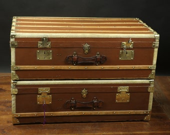 May include: Two vintage brown leather luggage trunks with brass hardware and leather handles. The trunks are stacked on top of each other.