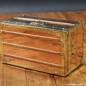 May include: A vintage-style wooden chest with brass accents and a lock. The chest is made of copper and has a wood interior. The chest is sitting on a wooden surface.