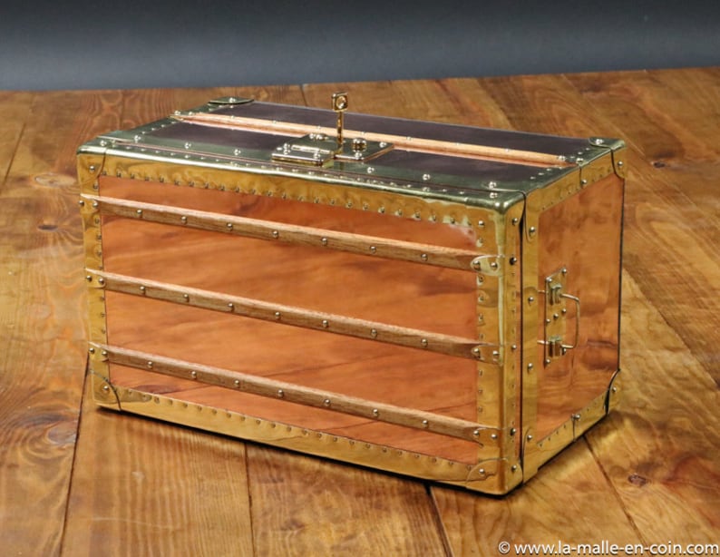 May include: A vintage-style wooden chest with brass accents and a lock. The chest is made of copper and has a wood interior. The chest is sitting on a wooden surface.
