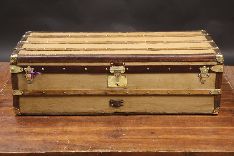May include: A vintage, tan canvas travel trunk with brown leather accents and brass hardware. The trunk has a hinged lid and a brass lock with a keyhole. The trunk is sitting on a wooden surface.