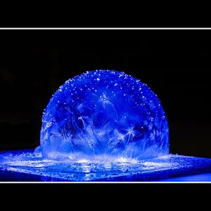 Op de afbeelding: Een gloeiend blauw, halfrond object met ingewikkelde, veervormige patronen en sprankelende highlights. Het object staat op een rechthoekige basis, alles tegen een diepzwarte achtergrond. Het geheel is een spel van licht en textuur.