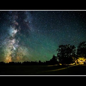 May include: A nighttime landscape featuring a vibrant display of the Milky Way. The sky is filled with countless stars, with a bright, colorful band of the galaxy dominating the scene. Silhouetted trees and a dark field are in the foreground.