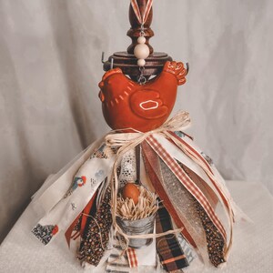 May include: A decorative wooden jewelry stand with a red ceramic chicken figurine. The stand is adorned with fabric ribbons and a small metal bucket with an egg. The stand is approximately 12 inches tall.