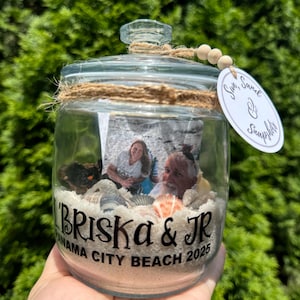 May include: A clear glass jar with a lid, filled with sand, seashells, and a photo of two people. The jar is decorated with twine and a tag that reads "Sun, Sand & Snapshots." The jar has the text "'Briska & JR Panama City Beach 2025" on the front.