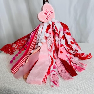 May include: A Valentine's Day tassel ornament with a pink heart-shaped charm that reads "BE MINE." The tassel features various pink and red ribbons, lace, and fabric strips. Small white pom-poms are interspersed throughout the design.