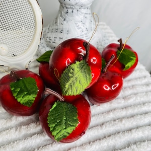 May include: Six glossy red apple ornaments with green fabric leaves and brown stems. Each apple has a gold string for hanging. The apples are arranged on a white, textured surface, suggesting a festive or decorative theme.