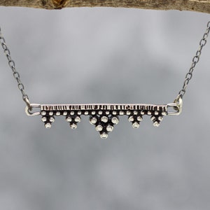 Puede incluir: Un collar de plata con una cadena delicada y un colgante geométrico. El colgante está hecho de pequeñas cuentas redondas dispuestas en un patrón.