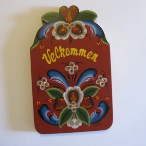 May include: A red wooden sign with a welcome message in yellow lettering and a floral design painted in blue, green, white, and brown. The sign says "Willkommen".