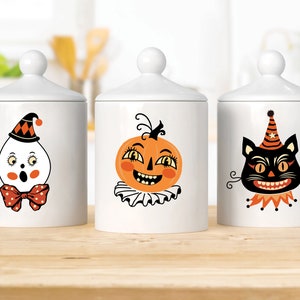 Peut inclure: Trois pots en c&eacute;ramique blanche avec des couvercles noirs et des illustrations sur le th&egrave;me d'Halloween. Les pots repr&eacute;sentent un fant&ocirc;me, une citrouille et un chat noir.