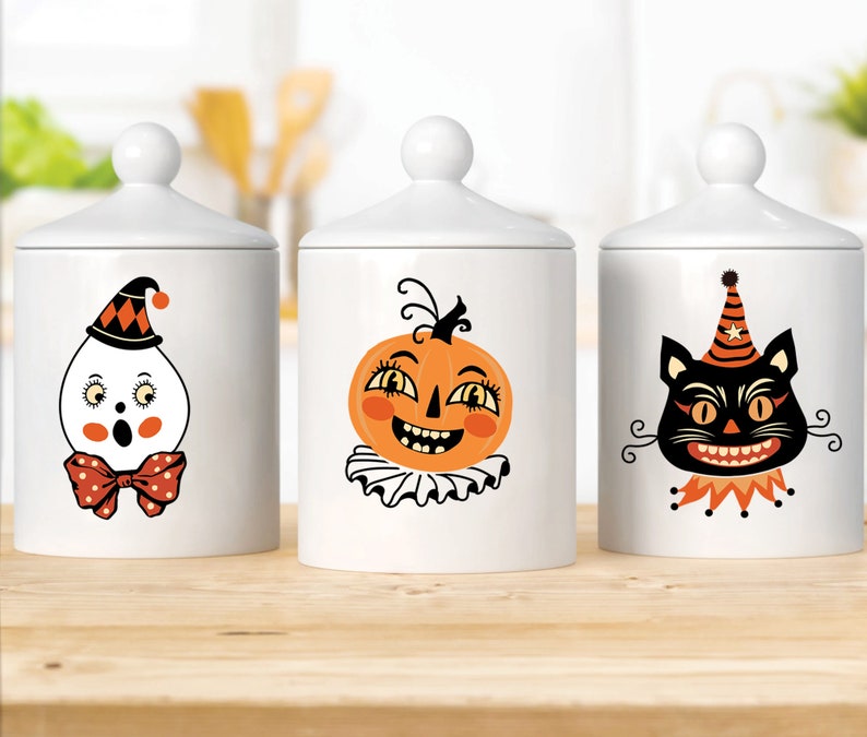 Peut inclure: Trois pots en c&eacute;ramique blanche avec des couvercles noirs et des illustrations sur le th&egrave;me d'Halloween. Les pots repr&eacute;sentent un fant&ocirc;me, une citrouille et un chat noir.