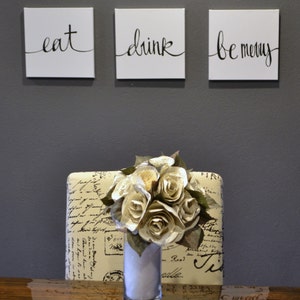 May include: Three square white canvases with black cursive text that reads "eat", "drink", and "be merry". Below, a bouquet of paper roses in a clear vase sits on a patterned surface. The background is a solid gray wall.