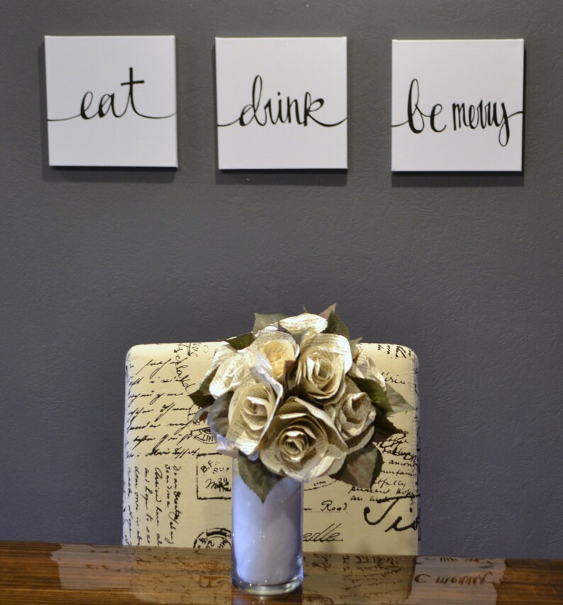 May include: Three square white canvases with black cursive text that reads "eat", "drink", and "be merry". Below, a bouquet of paper roses in a clear vase sits on a patterned surface. The background is a solid gray wall.