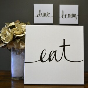May include: White canvas art with the word "eat" in black script. Two smaller square canvases above read "drink" and "be merry" in black script. A vase of paper roses is to the left.