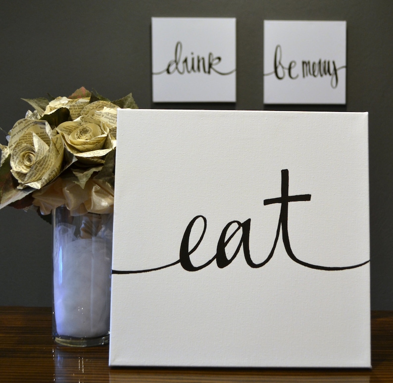 May include: White canvas art with the word "eat" in black script. Two smaller square canvases above read "drink" and "be merry" in black script. A vase of paper roses is to the left.
