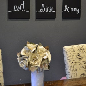 May include: Three black square wall art pieces with white cursive text that reads "eat", "drink", and "be merry". A bouquet of paper roses in a clear vase sits on a wooden surface.