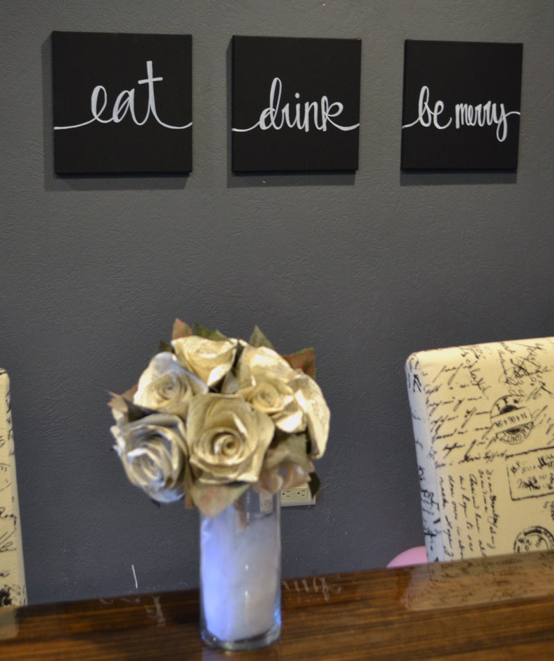May include: Three black square wall art pieces with white cursive text that reads "eat", "drink", and "be merry". A bouquet of paper roses in a clear vase sits on a wooden surface.