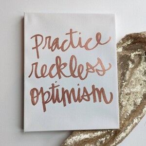 May include: White canvas art with the words "Practice reckless optimism" in copper-colored script. The canvas is rectangular and sits on a white surface with a gold sequined fabric in the background.