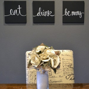 May include: Three black square wall art pieces with white cursive text that reads "eat", "drink", and "be merry". Below, a vase of paper flowers sits on a wooden surface, in front of a patterned chair.