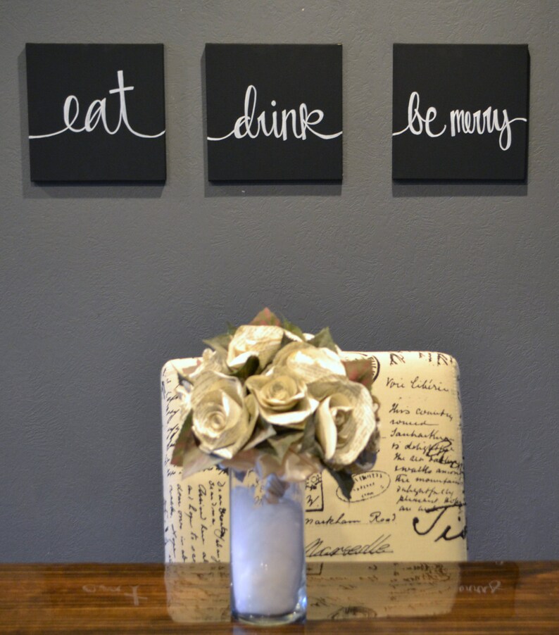 May include: Three black square wall art pieces with white cursive text that reads "eat", "drink", and "be merry". Below, a vase of paper flowers sits on a wooden surface, in front of a patterned chair.