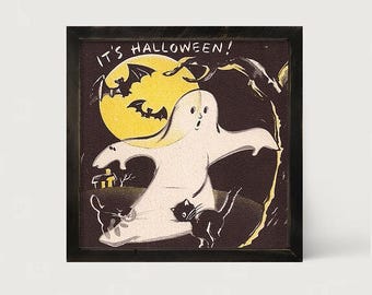 May include: A framed square print featuring a white ghost centered against a dark background with a large yellow moon, two black bats, and two black cats. The text IT'S HALLOWEEN! is printed in white at the top. The artwork has a textured finish.