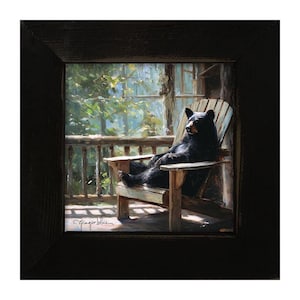 May include: A framed painting depicting a black bear sitting in a wooden Adirondack chair on a porch. The scene shows a forest background with sunlight filtering through the trees. The artist's signature, Ginger Blue, is visible in the bottom left corner.