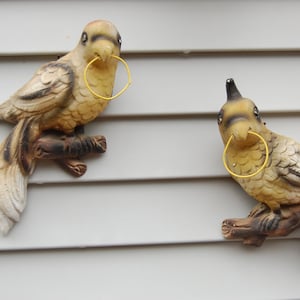 May include: Two ceramic bird figurines with yellow rings around their beaks, perched on branches, mounted on a white wall.