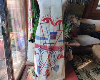May include: An off-white apron featuring a graphic print of gardening tools in red and blue. The design includes illustrations of shovels, rakes, and potted plants. The apron has a square neckline and thin straps, displayed on a mannequin torso.