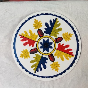 May include: A round, white decorative plate with a blue scalloped edge. The plate features a central design of a blue star surrounded by yellow, red, yellow, and blue leaves, and red acorns. The plate is likely made of metal.