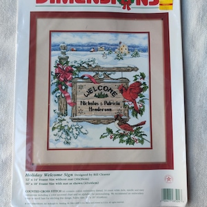 May include: A Dimensions counted cross-stitch kit for a holiday welcome sign. The framed artwork features a winter scene with a welcome sign, cardinals, and the names Nicholas & Patricia Henderson. The frame size is 16 x 18 inches with a mat.