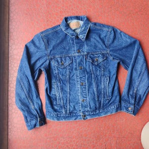 May include: A classic blue denim jacket with a collared neckline, button-down front, and multiple pockets. The jacket has long sleeves and a vintage look. The denim is a medium wash, and the jacket appears to be in good condition.