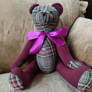 May include: A handmade teddy bear with a plaid body in shades of gray, burgundy, and red. The bear has burgundy arms, legs, and ears, and a bright purple bow. A button is sewn on the chest.
