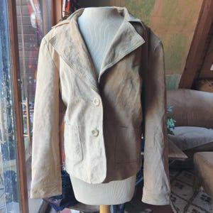 May include: A light beige suede jacket with a button closure and two pockets. The jacket has a collar and long sleeves.